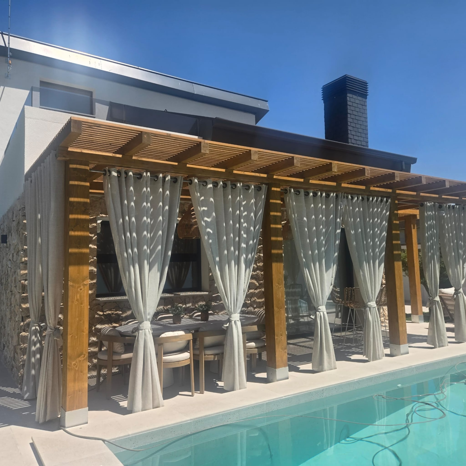 Pérgola de madera con cortinas junto a la piscina de la villa
