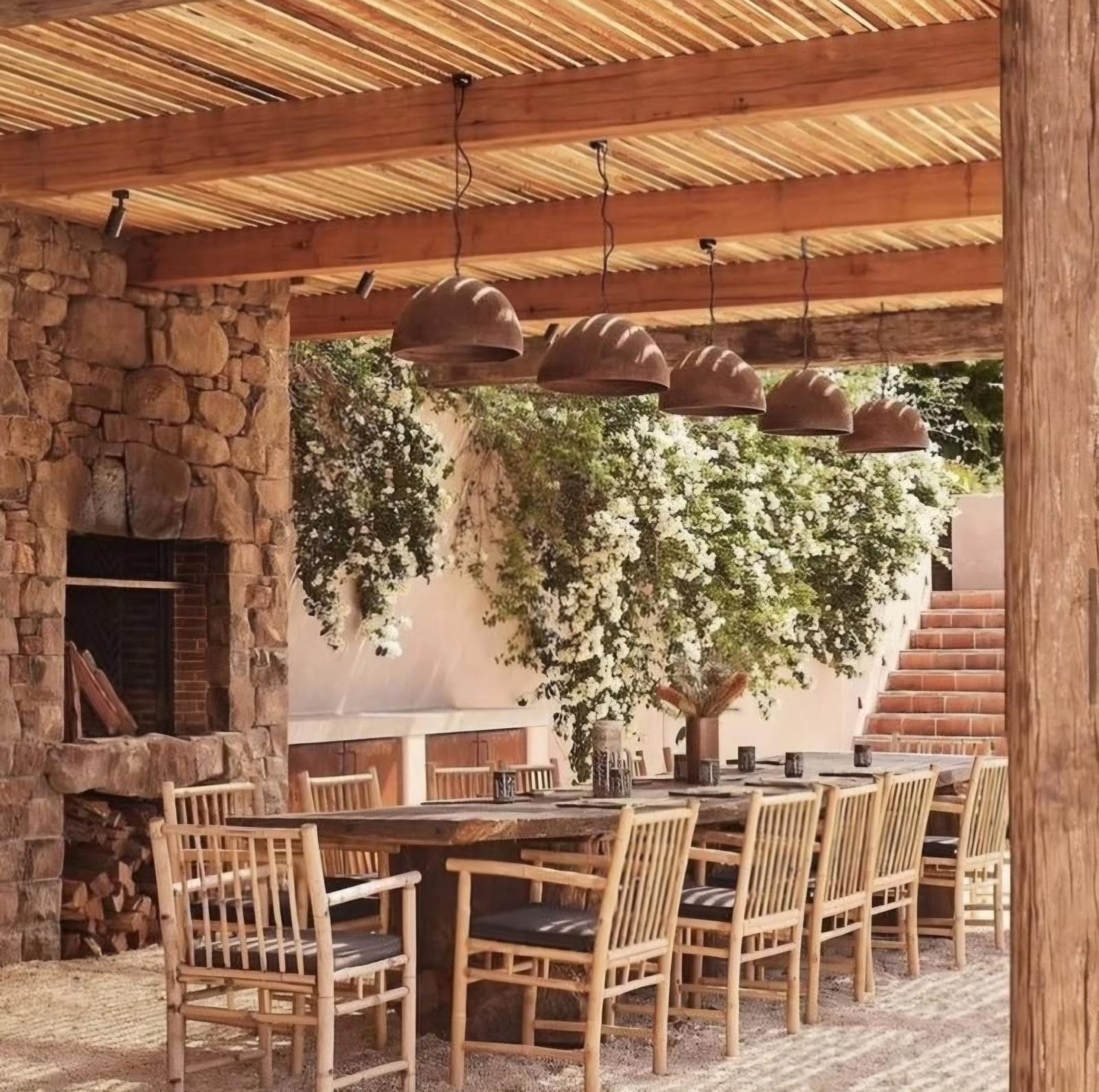 Mesa larga de madera bajo pérgola con lámparas de barro y muro de piedra